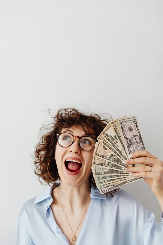 Joyful woman with curly hair and glasses holding US dollar bills excitedly.
